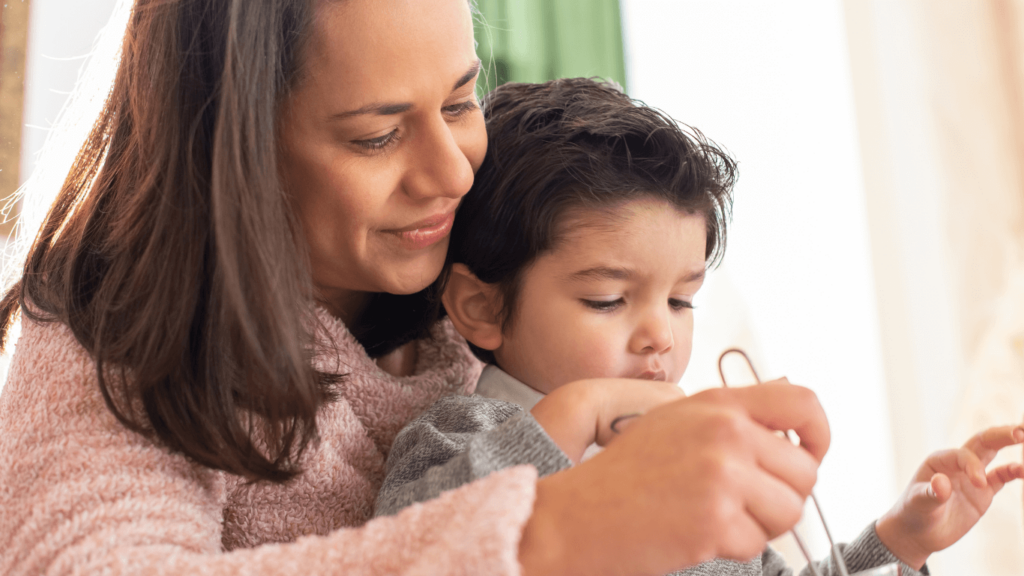 A woman lovingly helps a young child eat, gently guiding the child’s hand with MPS support. Both are seated closely together in a warm, softly lit room.