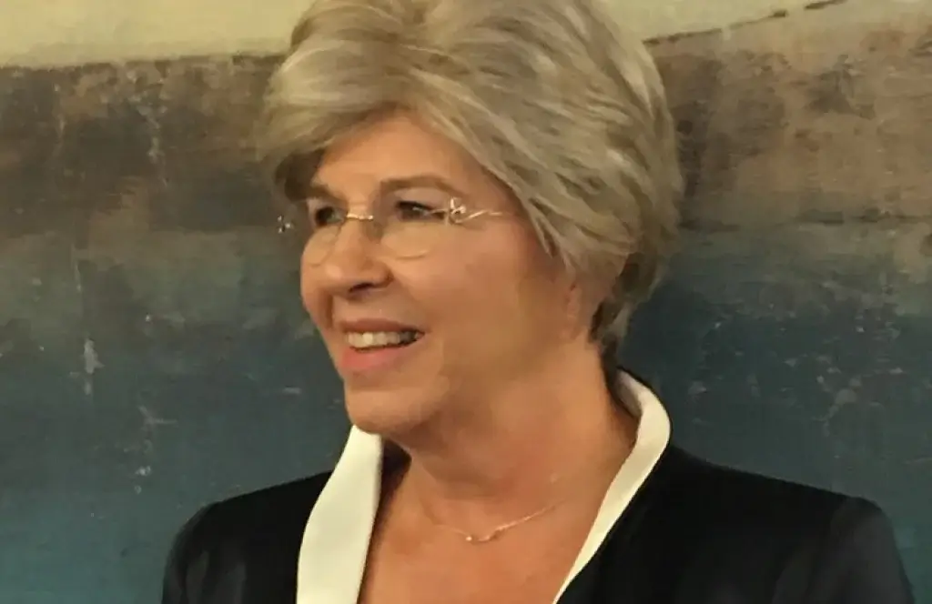 A smiling older woman with short, light gray hair and glasses, wearing a dark jacket with a white collar, stands in front of a textured, blue-and-brown background.